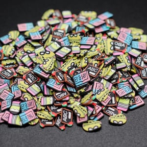 May include: A large pile of colorful, small, plastic, book-shaped charms with various designs and text, including "Eraser", "ABC", and "1926".