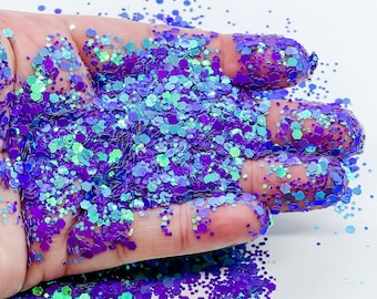 May include: A close-up of a hand holding a pile of iridescent purple and teal glitter. The glitter is a mix of different sizes and shapes, and it catches the light beautifully.