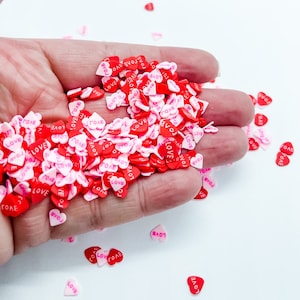 May include: A hand holding a large pile of small pink and red heart-shaped sprinkles with the word "LOVE" written on them.