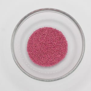 May include: A clear glass bowl filled with a large cluster of small, round, pink beads. The beads are densely packed together, forming a circular shape within the bowl. The bowl sits on a white surface.