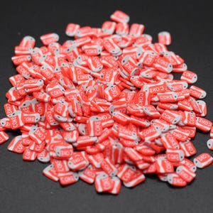 May include: A pile of miniature red and white Coke can charms. Each charm features the word "COKE" in white lettering on a red background, with a silver top and bottom. The charms are scattered on a dark surface.