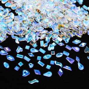 May include: A collection of iridescent, multi-faceted rhinestones in various shapes, including squares, rectangles, and teardrops. The rhinestones have a clear, blue, and purple color scheme, with a shimmering, reflective quality. They are scattered on a black surface.