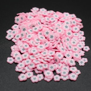 May include: A large pile of pink flower-shaped sprinkles with black centers. The sprinkles are scattered on a black surface.