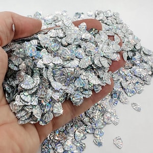 May include: A large pile of iridescent silver seashell-shaped glitter. The glitter is scattered on a white surface and a hand is holding a portion of the glitter.