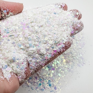 May include: White iridescent glitter with a mix of small and large glitter pieces. The glitter is in a hand, and some is spilling out onto a white surface.