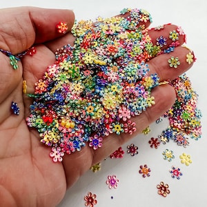 May include: A large pile of iridescent rainbow flower-shaped sequins in a hand. The sequins are scattered on a white surface.