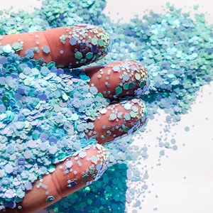 May include: A hand holding a pile of iridescent blue and green glitter. The glitter is scattered on a white surface.