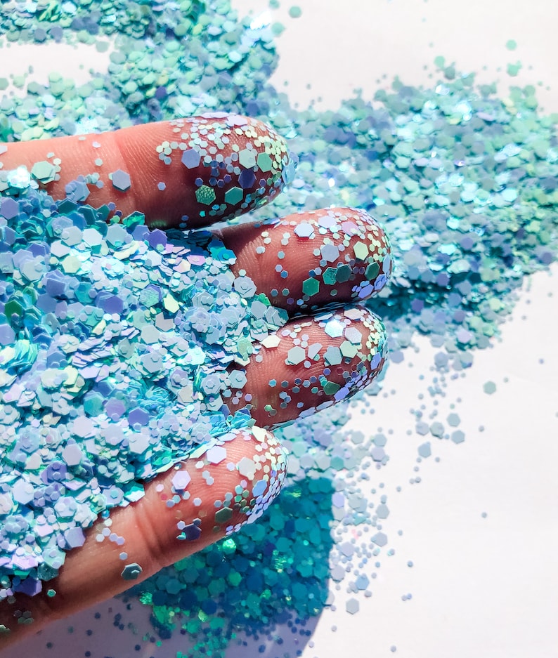 May include: A hand holding a pile of iridescent blue and green glitter. The glitter is scattered on a white surface.