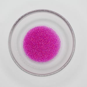 May include: A clear glass bowl filled with a vibrant cluster of small, magenta-colored beads. The beads are densely packed, forming a circular shape within the bowl. The bowl sits on a plain white surface, creating a contrast with the bright color of the beads.
