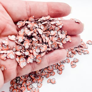 May include: A hand holding a large amount of small, orange bear face sprinkles. The sprinkles are scattered on a white surface.