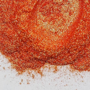 May include: Close-up of a pile of shimmering, orange-gold glitter. The glitter is densely packed, creating a textured surface with a metallic sheen. The glitter is scattered on a white surface.