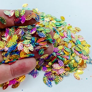 May include: A large pile of iridescent, multi-colored leaf-shaped sequins. The sequins are scattered on a white surface and are being held by a hand.