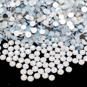 May include: A collection of iridescent, round-cut rhinestones. The rhinestones have a flat, silver backing and a slightly domed, opalescent top, creating a shimmering effect. The gems are scattered across a black surface.