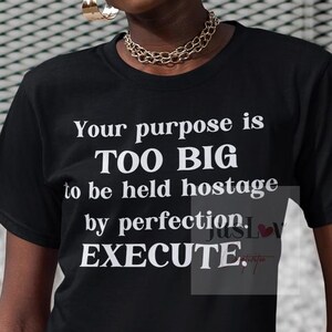 May include: Black t-shirt with white text: "Your purpose is TOO BIG to be held hostage by perfection. EXECUTE." The model is wearing glasses, gold jewelry, and blue jeans. The background is a gray, textured wall.