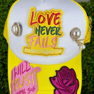 Può includere: Cappellino da camionista giallo e bianco con la scritta "LOVE NEVER FAILS" e "I WILL TRUST". Il cappellino presenta una toppa a forma di rosa rosa e dettagli di perle. Il cappellino è decorato con spille color oro.