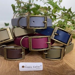 May include: A stack of six colorful leather dog collars with brass buckles. The collars are in shades of gray, beige, navy blue, burgundy, brown, and olive green. The collars are arranged in a stack with the buckles facing the camera. The text "The Happy Collar Company" is visible at the bottom of the image.