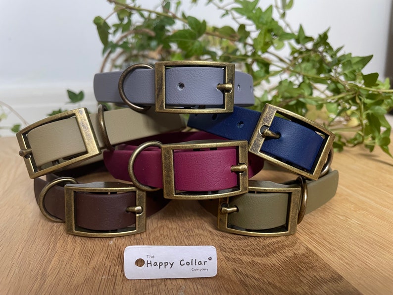 May include: A stack of six colorful leather dog collars with brass buckles. The collars are in shades of gray, beige, navy blue, burgundy, brown, and olive green. The collars are arranged in a stack with the buckles facing the camera. The text "The Happy Collar Company" is visible at the bottom of the image.