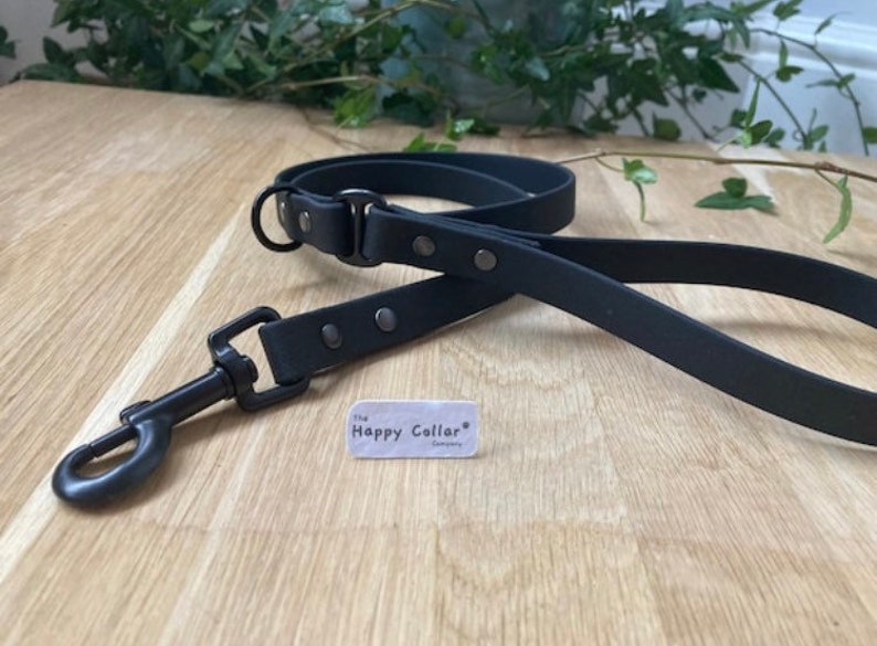 May include: Black leather dog leash with a metal clasp and silver accents. The leash is on a wooden surface with a small white tag that says "The Happy Collar Company".