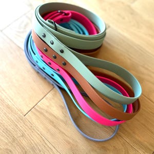 May include: A selection of colourful belts in shades of pink, blue, brown, and green, arranged on a wooden surface. Some belts feature metal stud details. The belts are coiled and layered, displaying a range of vibrant colours.