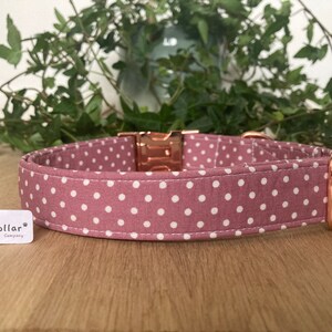 May include: A pink dog collar with white polka dots and rose gold hardware. The collar is made of fabric and has a buckle closure. The collar is labeled "The Happy Collar Company."