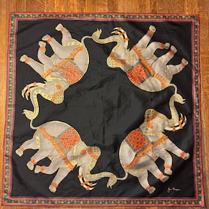 May include: Square black scarf featuring four grey elephants with orange and gold detailing in a repeating pattern. The scarf has a decorative border with teal, orange, and gold details. The scarf is made of a silky material.