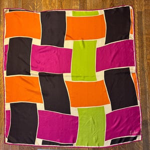 May include: A square silk scarf with a vibrant geometric design. It features large blocks of orange, black, fuchsia, and green, framed by off-white borders. The scarf has a pink border and the word "Echo" is printed in the corner.