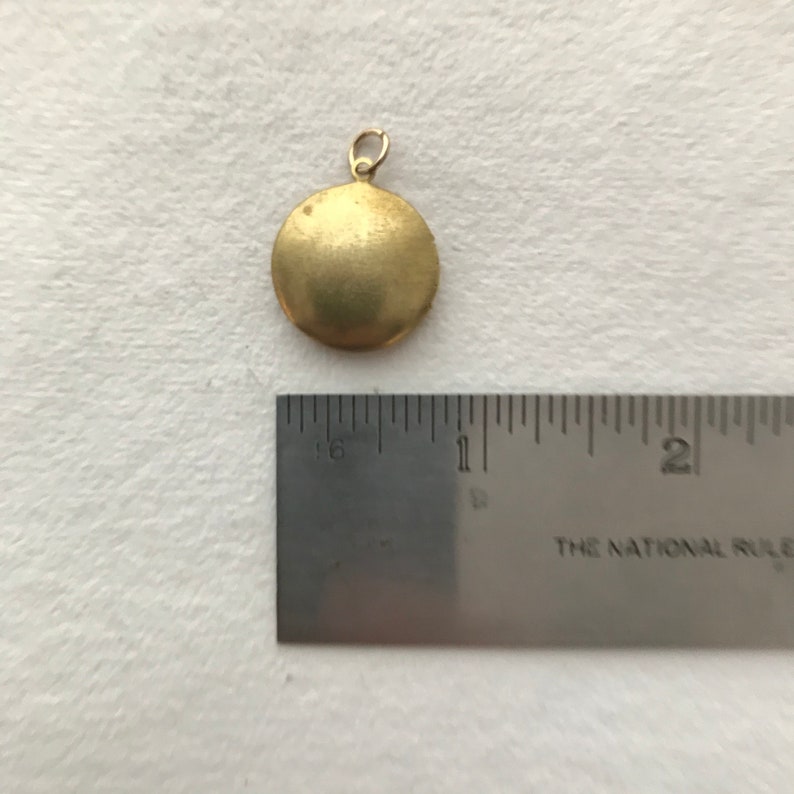 May include: A gold-colored round locket pendant with a smooth surface. The pendant is about 1 inch in diameter and is shown next to a ruler.