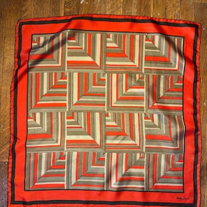 Peut inclure: Un foulard carré avec un motif géométrique de lignes angulaires répétitives orange, grises et blanches. Le foulard présente une large bordure orange avec un fin liseré noir. Un ruban à mesurer indique une largeur d'environ 58 cm.