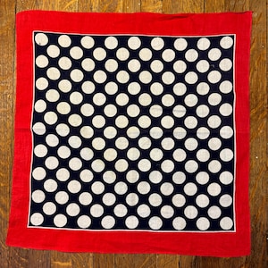 Könnte beinhalten: Ein quadratisches Bandana mit rotem Rand und marineblauem Zentrum mit weißen Polka-Dots. Der Stoff scheint Baumwolle oder ein ähnliches Material zu sein. Das Bandana liegt auf einer Holzoberfläche. Das Design ist ein klassisches Polka-Dot-Muster.