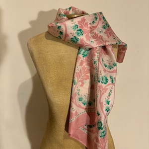 Puede incluir: Una bufanda rosa y blanca con un estampado floral y un borde rosa está drapeada sobre un maniquí. La bufanda presenta un diseño repetido de hojas rosas y flores turquesas.