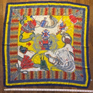 May include: A square silk scarf featuring a yellow centre with a central crest, red and blue equestrian-themed illustrations, and a decorative border of red and grey patterns. A white measuring tape shows the scarf is approximately 89 cm wide.