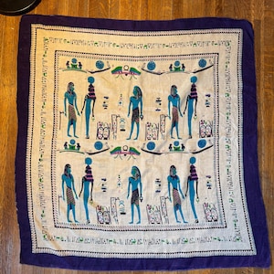 May include: A square scarf with an Egyptian theme. The scarf features a cream-coloured centre with blue and teal hieroglyphic figures and symbols. The border is a deep purple. The scarf is lying on a wooden surface.