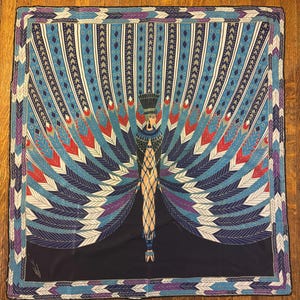 May include: A square silk scarf featuring a central figure in an Egyptian-inspired headdress and attire, surrounded by a large, fan-like display of blue, red, and purple feathers. A white measuring tape shows the scarf is 86 cm wide.