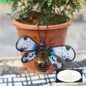 May include: A handcrafted metal butterfly sculpture with blue and white wings, a black body, and a glass orb abdomen. The sculpture is placed in front of a terracotta pot with a green plant.