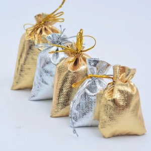 May include: Four small drawstring bags made of shimmery fabric. Two bags are gold and two are silver. The bags are lined up in a row on a white surface.