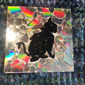 May include: A black cat silhouette is created using pieces of iridescent, shattered compact discs. The background is a light gray, and the cat is facing the right side of the image.