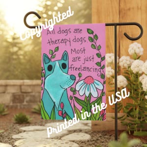 May include: A pink garden flag featuring a blue dog illustration and the text "All dogs are therapy dogs. Most are just freelancing." The flag is printed with the text "Printed in the USA".