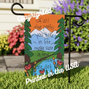 May include: A garden flag with a painted landscape scene. The flag features an orange sky, white mountains, green trees, and a blue river. Text on the flag reads "of all the paths you take in life, make sure a few of them are dirt."