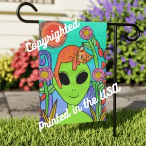 May include: A vibrant garden flag depicting a green alien with an orange cat resting on its head. The flag includes a sun, flowers, and the text "Copyrighted" and "Printed in the USA".