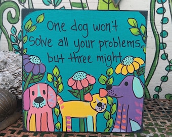 May include: A square wooden sign with a teal background featuring the hand-painted text One dog won't solve all your problems, but three might. The design includes three colorful dogs in pink, yellow, and purple, surrounded by flowers and green vines.