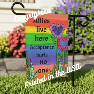 May include: A rainbow-colored garden flag with the text "Allies live here Acceptance hurts no one printed in the USA" and two purple flowers with hearts in the center.