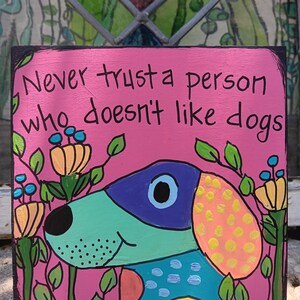 May include: A pink wooden sign with the text "Never trust a person who doesn't like dogs." The sign features a colorful cartoon dog with a blue and purple head, a teal body, and an orange ear with yellow spots. Green leaves and flowers surround the dog.