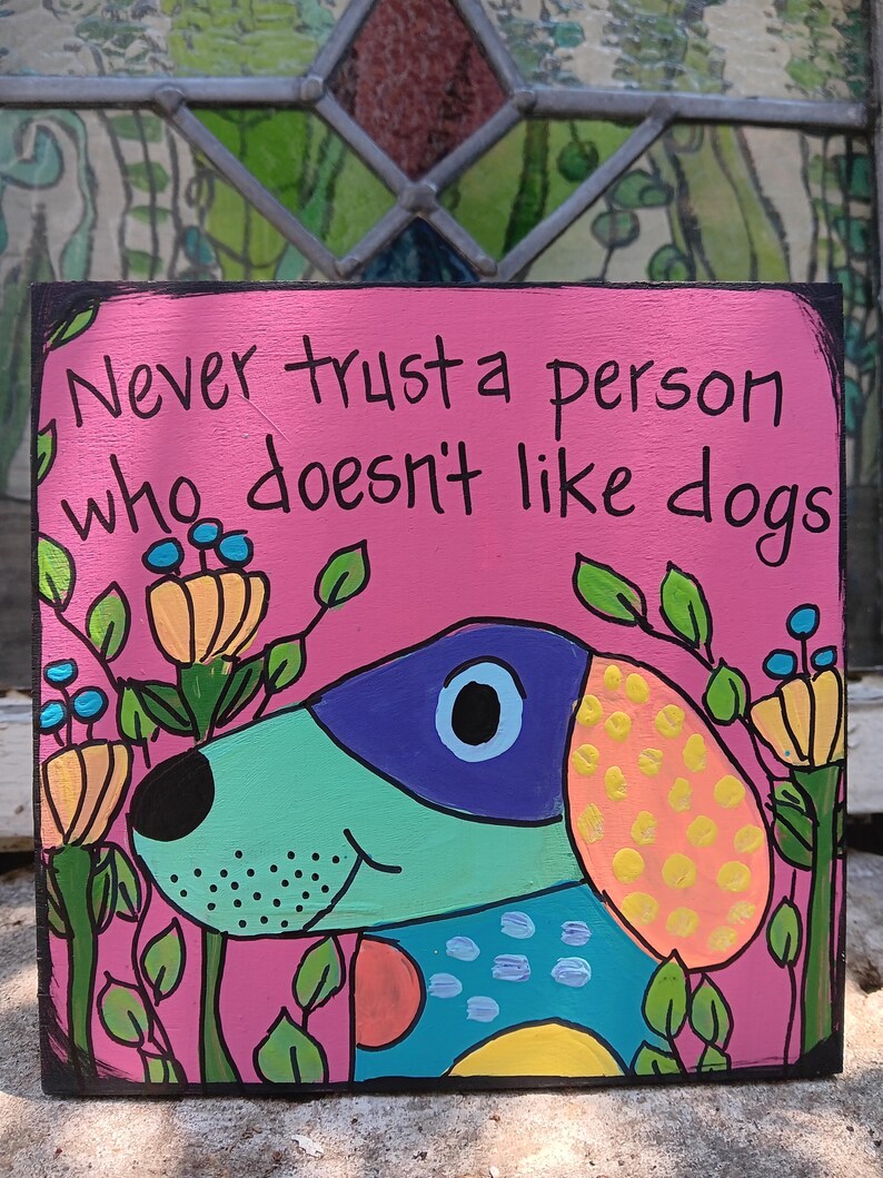 May include: A pink wooden sign with the text "Never trust a person who doesn't like dogs." The sign features a colorful cartoon dog with a blue and purple head, a teal body, and an orange ear with yellow spots. Green leaves and flowers surround the dog.
