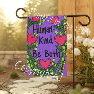May include: A purple garden flag with a green leafy border and pink hearts. The text on the flag reads "Human-Kind Be Both".