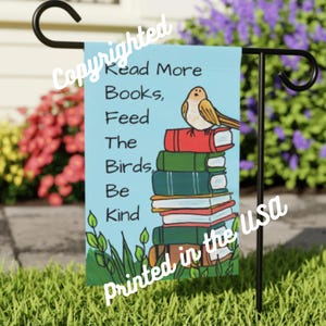 May include: A garden flag featuring the text "Read More Books, Feed The Birds, Be Kind" on a light blue backdrop. A bird illustration sits atop a stack of books. The flag is printed in the USA.