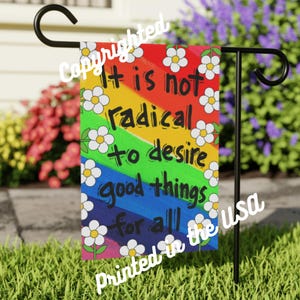 May include: A garden flag with a rainbow background and the words "It is not radical to desire good things for all." White daisies with yellow centers are scattered across the flag. The flag is printed in the USA.
