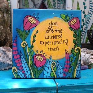 May include: A square canvas painting featuring a bright yellow sun against a blue background. The text "you are the universe experiencing itself" is written in black. Pink flowers with green stems and blue leaves surround the central sun.