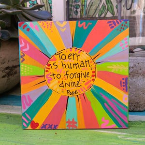 May include: Square, colorful artwork with a vibrant sunburst design in shades of orange, yellow, pink, and teal. The artwork features the quote "To err is human, to forgive, divine." by Pope, written in black. A heart and hashtag are included.