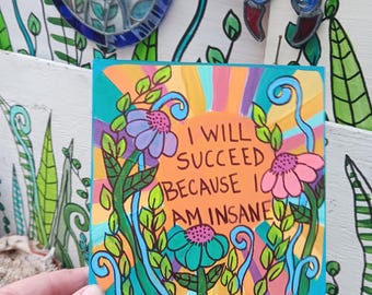 May include: A square art print featuring a vibrant sunburst background in shades of orange, yellow, and pink. The text "I WILL SUCCEED BECAUSE I AM INSANE" is written in black in the center, surrounded by colorful hand-drawn flowers and green vines.