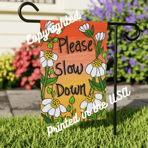 May include: A garden flag featuring the words "Please Slow Down" in black script, framed by white daisies and green foliage. The background fades from orange to yellow. The flag is mounted on a black metal stand.